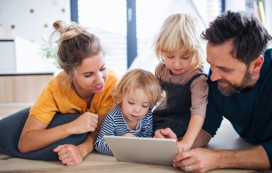 Front view of young family with two small children indoors in bedroom, using tablet.